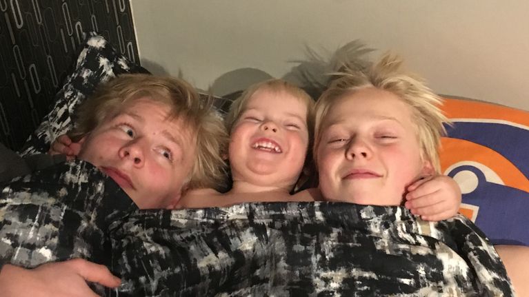 Kolme veljestä makaa peiton alla sängyssä ja hymyilee iloisesti.