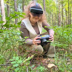 Woman kneeling in the forest wearing a goggle headset and holding a spectral imaging camera by a mushroom on the forest floor.