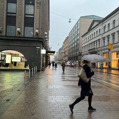 A fairly empty city street on a rainy day, with a few people walking on the sidewalk, a woman in the foreground is holding an umbrella.