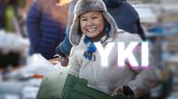 Nainen torilla kantaa laatikkoa ja hymyilee, kuvan päällä YKI-teksti.
