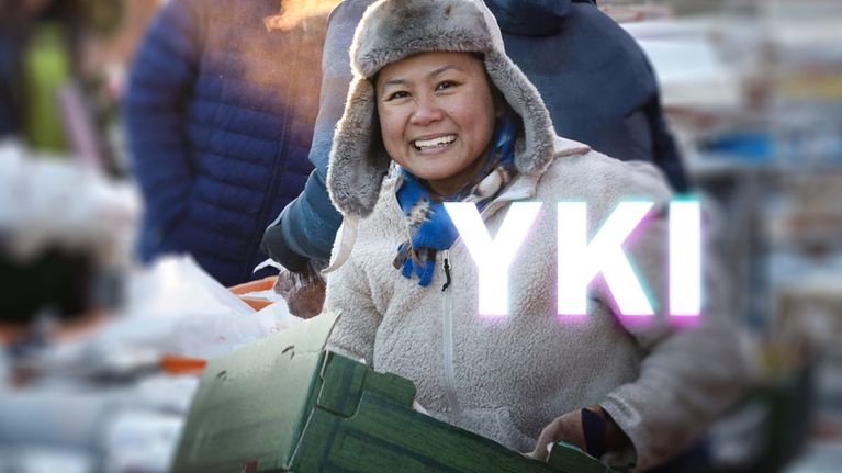 Nainen torilla kantaa laatikkoa ja hymyilee, kuvan päällä YKI-teksti.