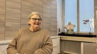 Harmaahiuksinen, silmälasipäinen, beigeen mekkoon puheutunut vanhempi naishenkilö istuu toimistotilassa, taustalla ikkunalaudalla naisen torso ja orkidea.