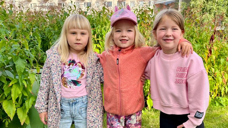 Tre små flickor står bredvid varandra med armarna om varandras axlar. Bakom dem grönt gräs och buskar, längre bort ett hus.