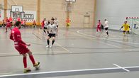 Futsal-peli käynnissä sisätiloissa: punaisiin ja valkoisiin pukeutuneet pelaajat liikkuvat kentällä. Taustalla näkyy yleisöä ja maalivahti valmiina maalilla.