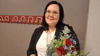 Nainen seisoo kukkapaketti kainalossa käytävällä ja katsoo vakavana kameraan.