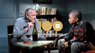 Harmaapäinen tupakoiva mies keskustelee nuoren tummaihoisen pojan kanssa pöydän ääressä. Kuvassa logo Yle 100 toive-elokuvaa.