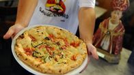Mies ojentaa lautasta, jolla pizza, vieressään Napolin suojeluspyhimystä San Gennaroa esittävä pysti.