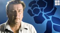 Aki Kaurismäki poseeraa kameralle, taustalla suuri tyylitelty ruusu sinisessä julisteessa.