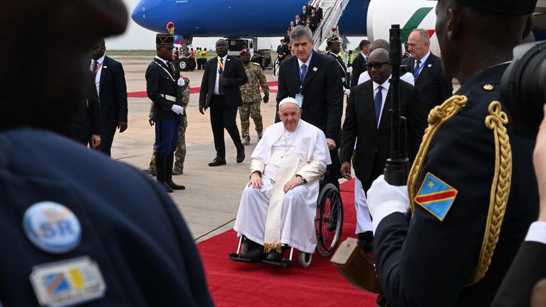 Påve Franciskus i rullstol på röda mattan i Kinshasa. Han mottas av ett hedersgarde av kongolesiska soldater.