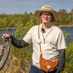Henkilö seisoo puisen veneen vieressä järven rannalla, taustalla vettä, metsää ja rakennus. Henkilöllä on lierihattu ja rintamerkki.
