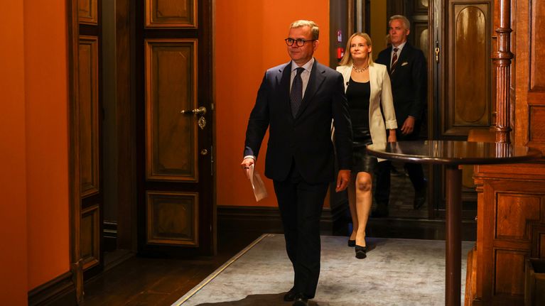 Kolme ministeria kävelee puupaneloituun, oranssisävyiseen huoneeseen, etualalla Petteri Orpo pitää paperia kädessään.