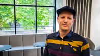 En ung man klädd i randig skjorta och keps står i en loungemiljö med stora fönster med träd utanför.