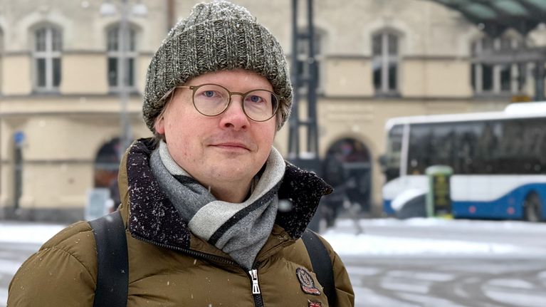Kuvassa silmälasipäinen mies pipo päässä katsoo kameraan. 
