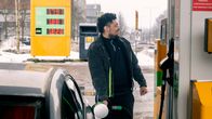 A man with dark hair, beard and jacket stands pumping petrol in his car in a snowy cityscape.