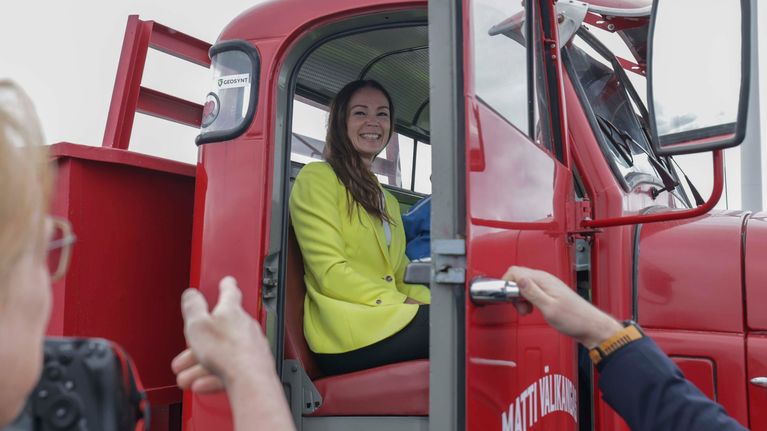 A woman with long dark hair and a yellow blazer sits smiling in a red lorry.