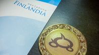Ruskealla pyödällä sinivalkoinen paperi, jossa lukee Sarjakuva-Finlandia. Vieressä kullattu mitali, jossa puhekuplia.