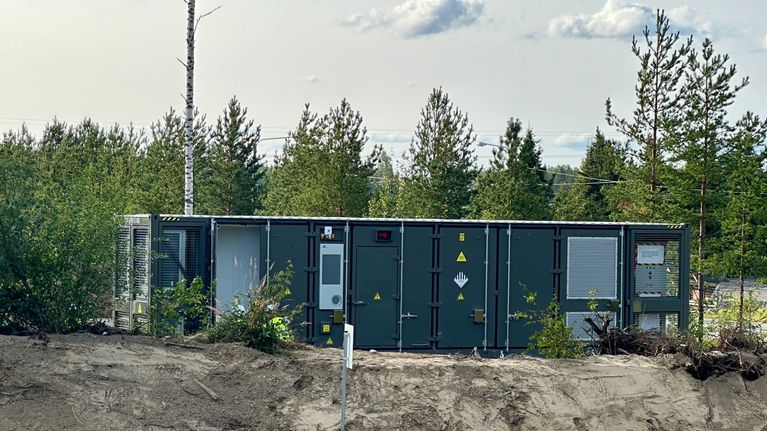 Vihreä akkukontti keskellä männikköä. Vieressä on hiekkapenger. Kontti on vihertävä.