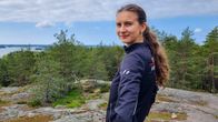 En person står på en höjd med utsikt över skog och sjö. Det är molnigt väder, men ändå ljust.