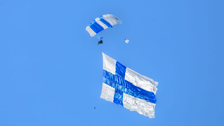A skydiver descending from the sky with a parachute waving a huge white and blue Finnish flag .