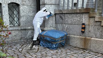 En person i vit skyddsdräkt undersöker ett blått objekt intill en stenmur och trappa utomhus.