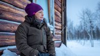 Nainen istuu lumihangella ja katselee kaukaisuuteen vanhan hirsitalon vierustalla. Taustalla lumista maisemaa ja koivuja.