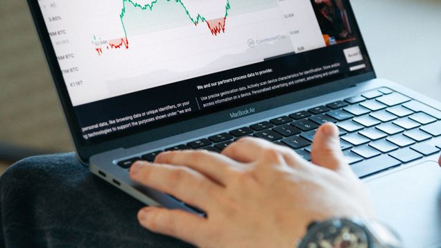 A hand on a laptop keyboard with the screen showing a stock graph.