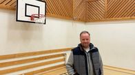 En man står en gymnastiksal med en korgborgställning bakom sig.
