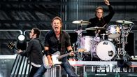 Bruce Springsteen haara-asennossa kitaransa kanssa ja laulaa. Takana kitaristi ja rumpali.