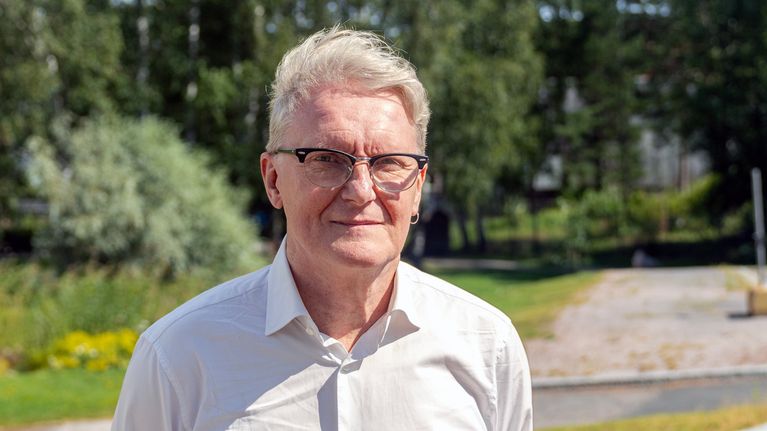 Mies jolla on vaalea paita , katsoo kameraan. Taustalla näkyy koivuja.