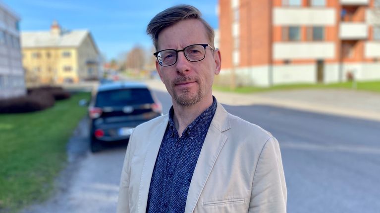En man i beige kavaj och blå skjorta och glasögon står ute i solen.