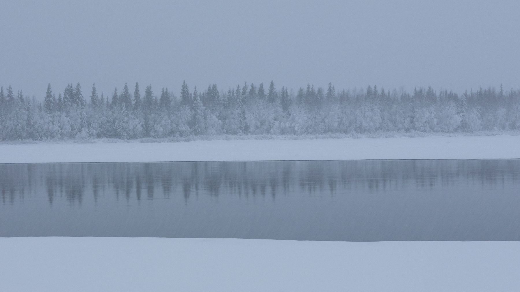 Talvinen maisema rauhallisen joen rannalla, jossa lumiset puut heijastuvat veden pintaan.