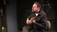 En man sitter på en stol i ett inspelningsrum och spelar gitarr, sladdar och ljudutrustning syns i bakgrunden.