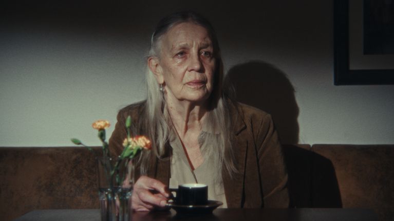 En äldre kvinna med långt grått hår sitter vid ett bord med en kopp kaffe och en vas med blommor framför sig.