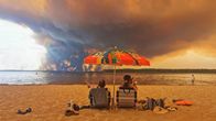 Two people sitting in beach chairs on a sandy beach, with a large cloud of smoke rising from the forest in the background. A colourful parasol shows the number "51".