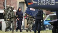 Militärklädda män leder en man i handfängsel, klädd i svart träningsoverall och stora hörlurar, mot ett flygplan på vars bakdel det står: Bundespolizei.