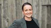 En glad kvinna i grå kavaj med armarna i sidan står framför en grå uthusvägg. 