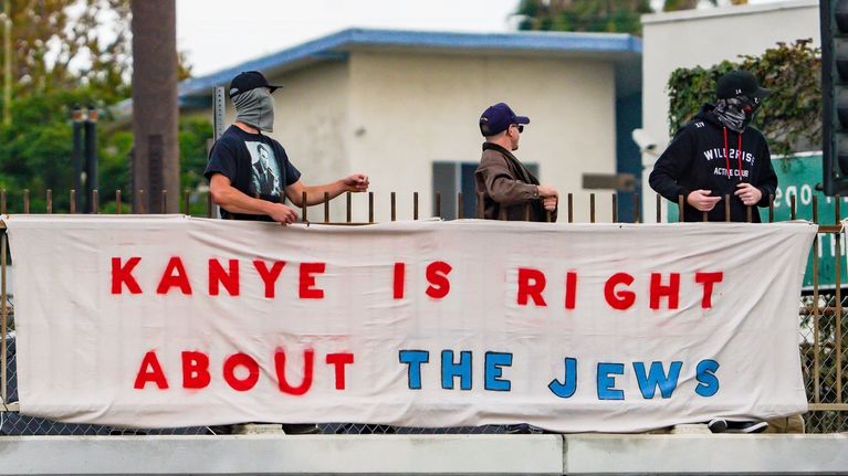 Kolme ihmistä, joista kaksi on peittänyt kasvonsa, pitää lakanaa, jossa lukee Kanye is right about the jews.