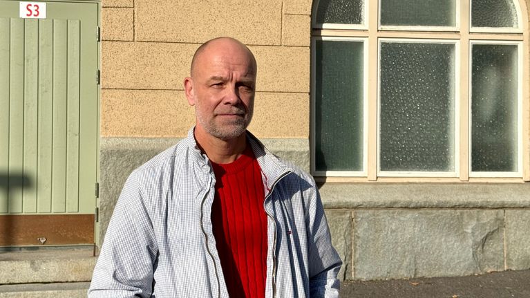 Henkilö seisoo rakennuksen edessä, pukeutuneena punaiseen neuleeseen ja vaaleaan takkiin. Taustalla näkyy vaaleanvihreä ovi ja ikkunoita.