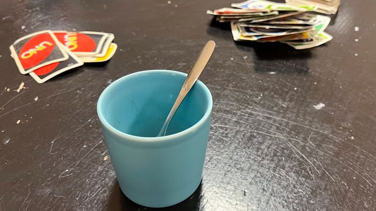 Turkoosi kahvikuppi on tummalla pöydällä, lusikka sojottaa mukista. Mukin molemmin puolin on kaksi pinoa Uno-pelikortteja.