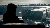 Maankaatokuljetus talvisessa auringonpaisteessa, jossa kuorma-auto kulkee lumisella tiellä. Etualalla näkyy tumma siluetti, taustalla talvinen maisema.