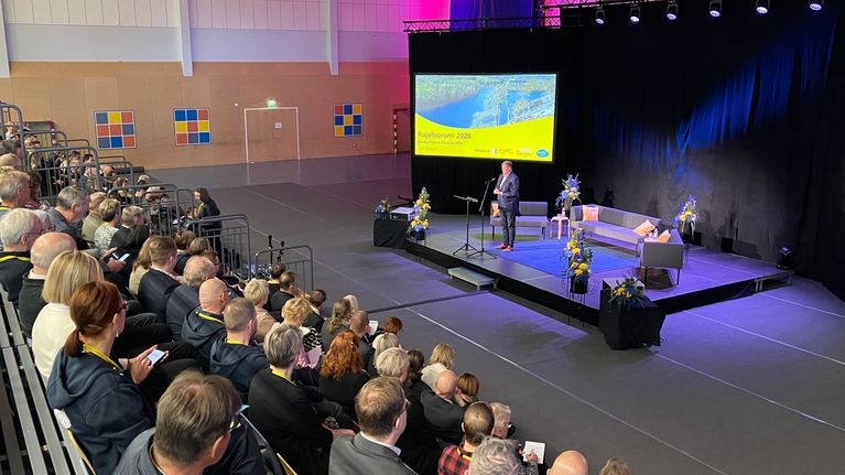 Kuntien edustajat seuraavat liikuntahallin lehtereillä lavalla puhuvaa Harri Bromania.