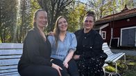 Kolme naista istuu valkoisella penkillä ulkona, taustalla punainen rakennus ja puita.