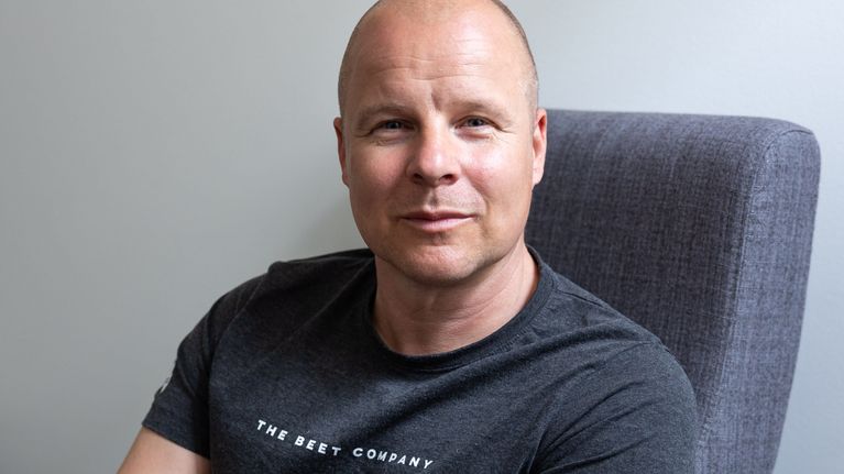En man sitter på en stol vid en grå vägg och tittar rakt in i kameran. 