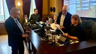 Flera personer står och sitter vid en upphöjd bänk i ett mötesrum med stora fönster; en stor skärm med tabell syns i bakgrunden.