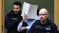 Kuvassa keskellä on mies, joka pitää kasvojensa edessä kansiota. Hänen molemmilla puolillaan seisoo poliisi.