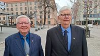 Två personer i kostym står utomhus på ett torg med byggnader i bakgrunden.