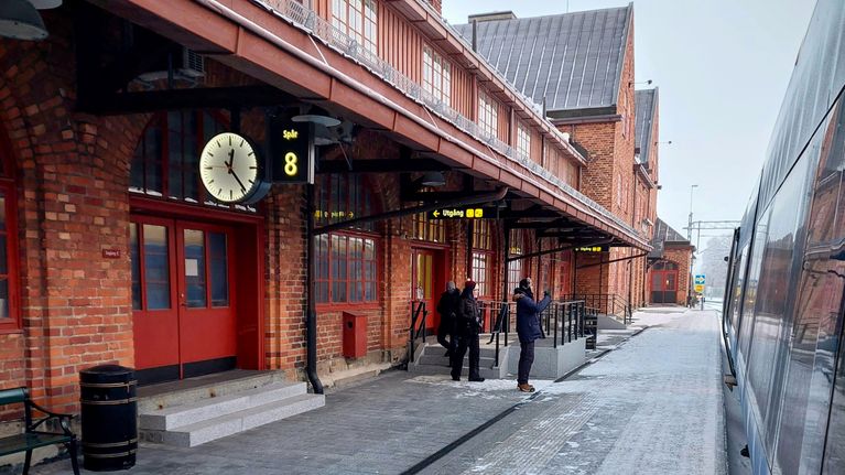 Three people on a platform with a red-brick station to the left and a silver train to the right.