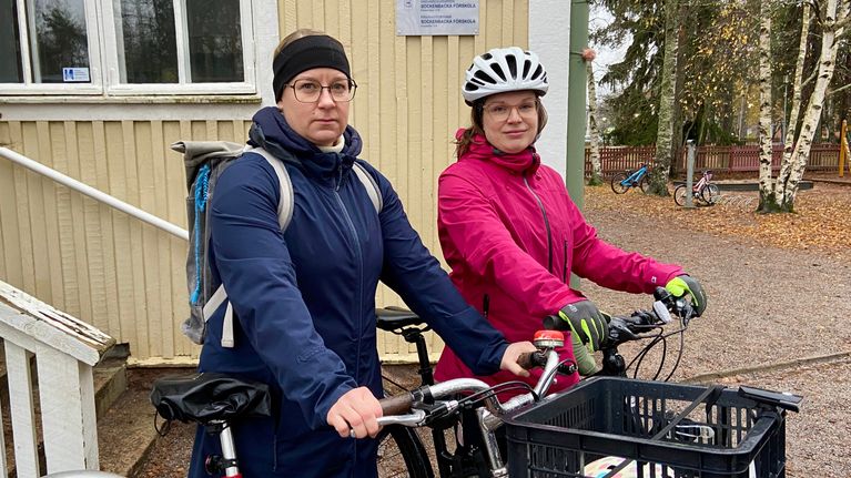 Två personer står med sina cyklar framför en byggnad. De är klädda för en cykeltur, och den ena personen har en cykelhjälm på sig.