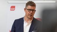 A man in short hair and glasses, Antti Lindtman, stands in front of wall displaying the SDP logo.