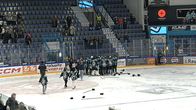 Musta ja turkoosipaitaiset Pelicans naiset joukkueen pelaajat ovat ringissa maalin ympärillä. Jäällä on mailoja ja varusteita. 